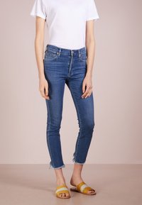 Jeans skinny de mezclilla azul con dobladillo deshilachado, combinados con sandalias de slide amarillas y grises, y una camiseta blanca ajustada.