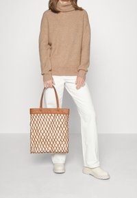 Gewebte tanfarbene Tragetasche mit einem Holzgriff, mit einem Rautenmuster und einem beigen Boden. Das Modell trägt einen beigen Pullover und weiße Hosen.