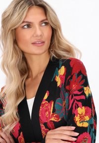 Donna con lunghi capelli biondi che indossa un cardigan nero con motivi floreali rossi, gialli e teal, con le braccia incrociate e che guarda leggermente di lato.
