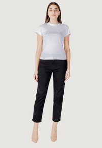 Witte t-shirt met korte mouwen met een zilveren hart en het "GUESS" logo, gecombineerd met zwarte strakke jeans en naaldhakken in huidskleur. Glad stofoppervlak.