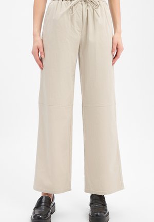 Beige broek met wijde pijpen, elastische taille en trekkoord, gedragen met zwarte glanzende loafers, zichtbaar van taille tot voeten.