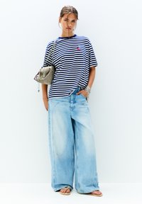 Stribet navy og hvid oversized t-shirt, lyseblå bredbenede jeans og en lille grå taske. Modellen bærer flade sandaler og minimalistisk smykker.