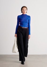 Top de gola alta azul com textura canelada, calças largas pretas, cinto preto, segurando uma bolsa de tote branca. Textura suave, design moderno, estilo minimalista.