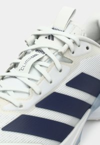 Chaussure de sport blanche avec une tige en mesh texturé, accents bleus et bandes bleu foncé, dotée de lacets ronds et d'une languette avec un onglet.