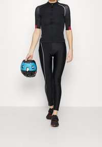 Personne portant une tenue de cyclisme noire et des chaussures noires, tenant un casque de vélo noir et bleu dans la main gauche, debout devant un fond blanc.