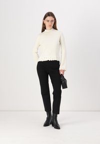 Pull à col roulé crème avec bord en dentelle en bas, associé à un pantalon noir sur mesure et des bottines, tenant un petit sac noir.
