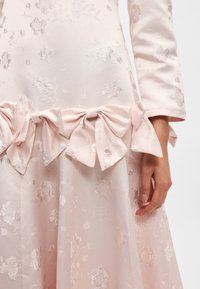 Ανοιχτό ροζ φόρεμα με λουλουδάτη υφή, διαθέτοντας μια μεγάλη λεπτομέρεια φιόγκου στη μέση και μακριά μανί�κια. Λείο ύφασμα με διακριτική γυαλάδα.