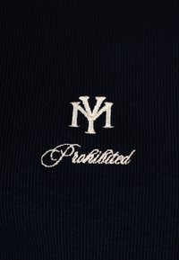 Logo NYM brodé en blanc au-dessus du mot « Interdit » en écriture cursive sur un tissu côtelé foncé.