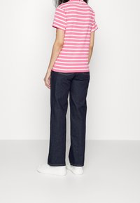 Žena v růžovobíle pruhovaném tričku s krátkým rukávem, tmavě modrých džínách a bílých teniskách, stojící zády k bílému pozadí.