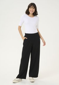 Pantaloni neri a gamba larga con una texture liscia e vita elastica, abbinati a un top bianco attillato a maniche corte e sneaker bianche.