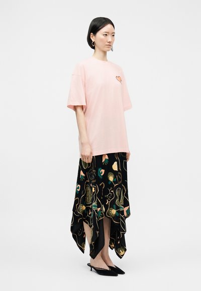 Γυναίκα με κοντά μαύρα μαλλιά, φοράει ροζ φαρδιά μπλούζα με καρδιά και μαύρη ασύμμετρη φλοράλ φούστα με μαύρα μυτερά παπούτσια.