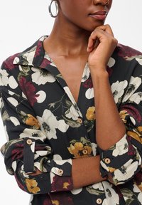 Femme à la peau foncée portant une chemise noir à fleurs avec des manches retroussées et des boucles d'oreilles en argent, reposant sa main sur son menton.