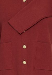 Burgundijas krāsa kardigāns ar teksturētu adījumu, ar pogu priekšpusi un zelta piederumiem, kā arī divām priekšējām kabatām. Mīksta auduma, ikdienišķs dizains.
