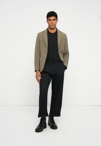 Khaki blazer met twee knopen, zwart shirt en zwarte wijde broek, gecombineerd met zwarte veterschoenen. Gladde texturen en strakke lijnen.