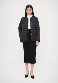 Μαύρο μπουφάν τύπου puffer με κουμπωτό μπροστινό μέρος, καπιτονέ σχέδιο και στρογγυλή λαιμόκοψη, συνδυασμένο με εφαρμοστή μαύρη φούστα και μαύρα παπούτσια.
