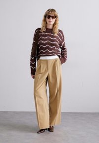 Brun strikket sweater med bølgemønster, cremefarvet top, beige vide bukser og brune hæle. Modellen bærer sorte solbriller og har en taske med.