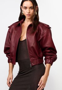 Burgundská kožená bomber bunda s rebrovaným lemom, zipsom a detailmi na pleciach. Hladká textúra a voľný strih.