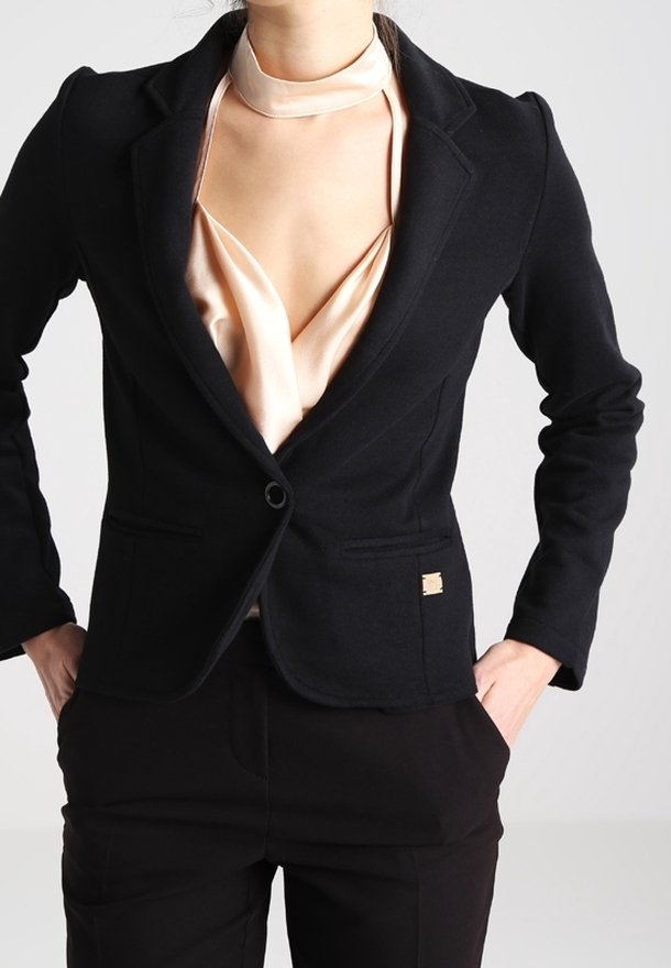 Zwarte fitted blazer met een tailored ontwerp, enkele knoopsluiting en een gouden accent op de zak. Satijnen beige top zichtbaar eronder.
