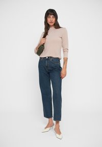 Top de manga larga beige combinado con jeans rectos azul oscuro y mule blancas. La modelo sostiene un bolso verde, mostrando un ajuste relajado.