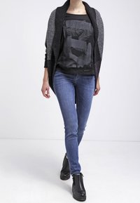 Kvinna som bär en svart mönstrad kofta, svart grafisk t-shirt, blå tajta jeans och svarta ankelstövlar går framåt på vit bakgrund.