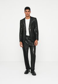 Zwart sequined pak met een getailleerde blazer en taps toelopende broek, gecombineerd met een wit T-shirt en zwarte schoenen. Strakke textuur en modern ontwerp.