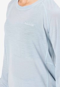 Světle modrá košile s dlouhým rukávem vyrobená z měkké, strukturované látky. Má kulatý výstřih a nenápadné logo vyšívané na hrudi.