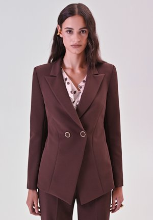 Brauner, doppelt geknöpfter Blazer mit Reverskragen, verziert mit goldenen Hardware-Details. Getragen über einer hellrosa Bluse mit Punkten. Glatte Stofftextur.