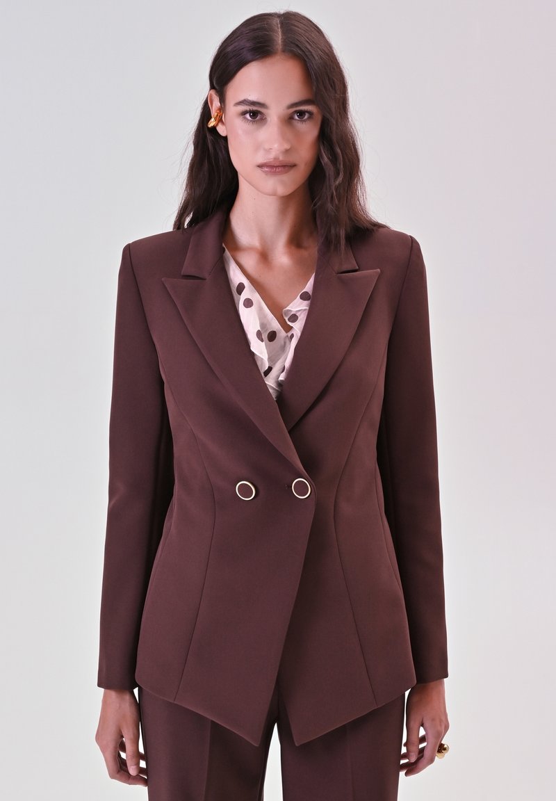 Blazer marrone doppiopetto con rever a punta, con dettagli in metallo dorato. Indossato sopra una blusa leggera rosa con pois. Texture del tessuto liscia.