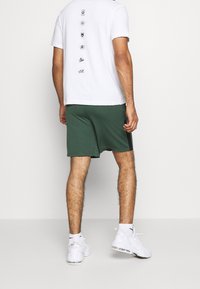 Pantalones cortos verdes hechos de una tela ligera, combinados con una camiseta blanca con estampados gráficos en la espalda. Combinados con zapatillas blancas.