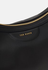 Sac en cuir noir avec une fermeture éclair dorée et un logo "Ted Baker" en relief sur une surface lisse. Design courbé notable et détails de couture.