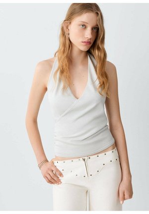 Jeune femme aux longs cheveux ondulés, portant un top cache-cœur blanc sans manches et un pantalon blanc taille haute avec fermeture éclair et détails cloutés.