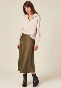 Maglione beige a maglia con colletto zip e bottoni ai polsini, abbinato a una gonna midi in satin verde oliva e stivaletti neri con tacco.