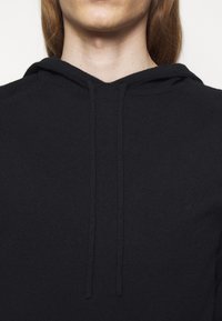 Frau mit einer schwarzen Strick-Hoodie mit Zugkordeln, die Nacken und oberen Brustbereich vor einem schlichten weißen Hintergrund zeigt.