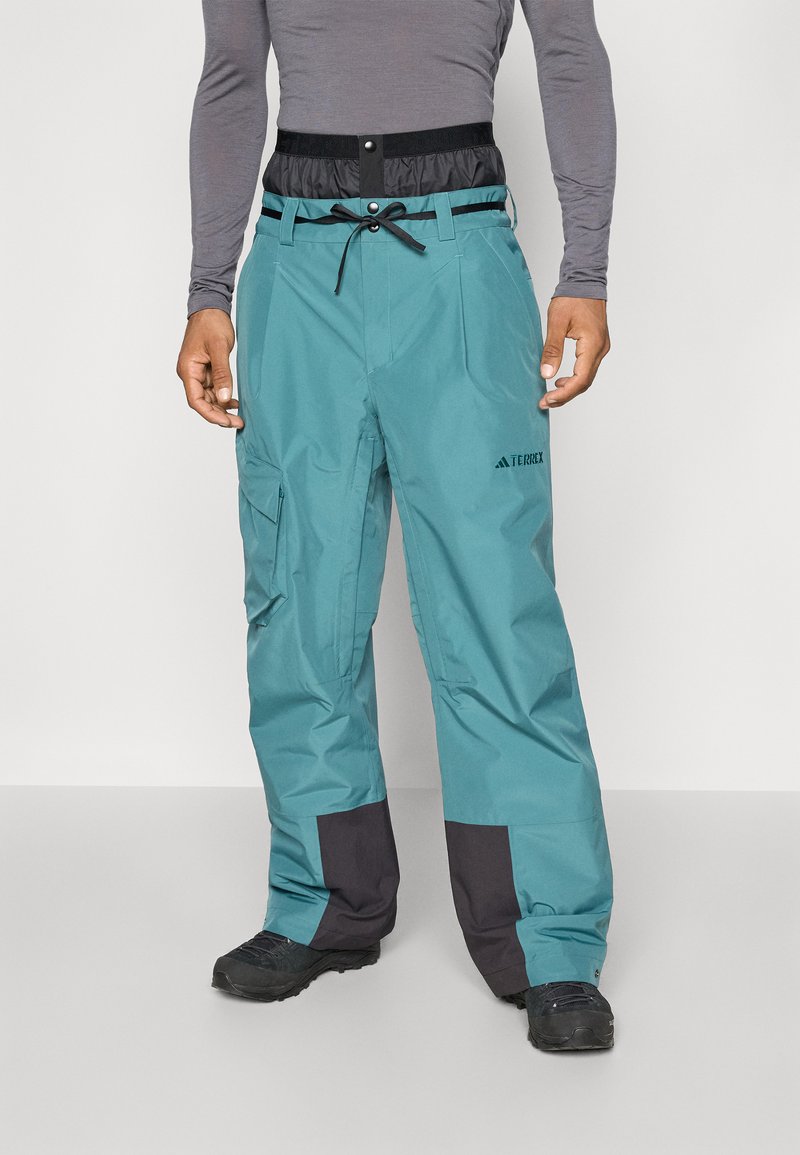 Hombre con pantalones deportivos azul verdoso y negros con bolsillo de carga y zapatos negros, de pie contra un fondo liso.