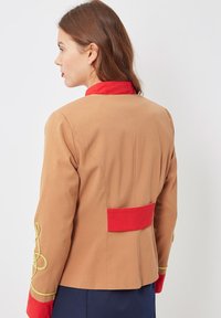 Mujer de perfil a la izquierda, vistiendo una chaqueta color beige con cuello rojo, tirantes rojos en la espalda y bordados en oro en las mangas, con fondo blanco.