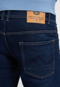 Jeans de mezclilla en azul oscuro con una tela texturizada. Presentan una etiqueta de cuero marrón con el logo "Petrol" y acentos de costura en los bolsillos traseros.