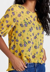 Haut jaune à manches courtes avec un motif floral comprenant des fleurs violettes et des feuilles vertes. Fabriqué dans un tissu léger avec une coupe décontractée.