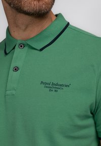 Zelené polo tričko s texturovaným povrchem, námořnický detail na límci, tři knoflíky a vyšívané logo "Petrol Industries" na hrudi.