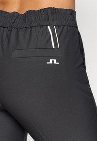 Čierne športové nohavice s textúrovým povrchom, elastickým pásom, bočnými vreckami a bielymi pruhmi na stranách. Obsahujú detail s logom.