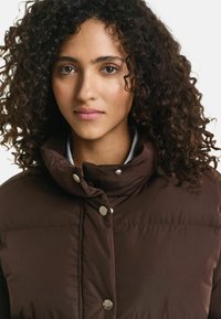 Bruine pufferjack met een hoge kraag, voorzien van zilveren drukknoopjes en een gladde textuur. Model heeft krullend haar en een neutrale uitdrukking.