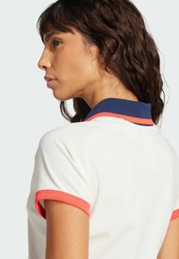 Biely polo tričko s námorníckym golierom a koralovými detailmi na rukávoch. Tkanina vyzerá hladko a vo voľnejšom strihu.