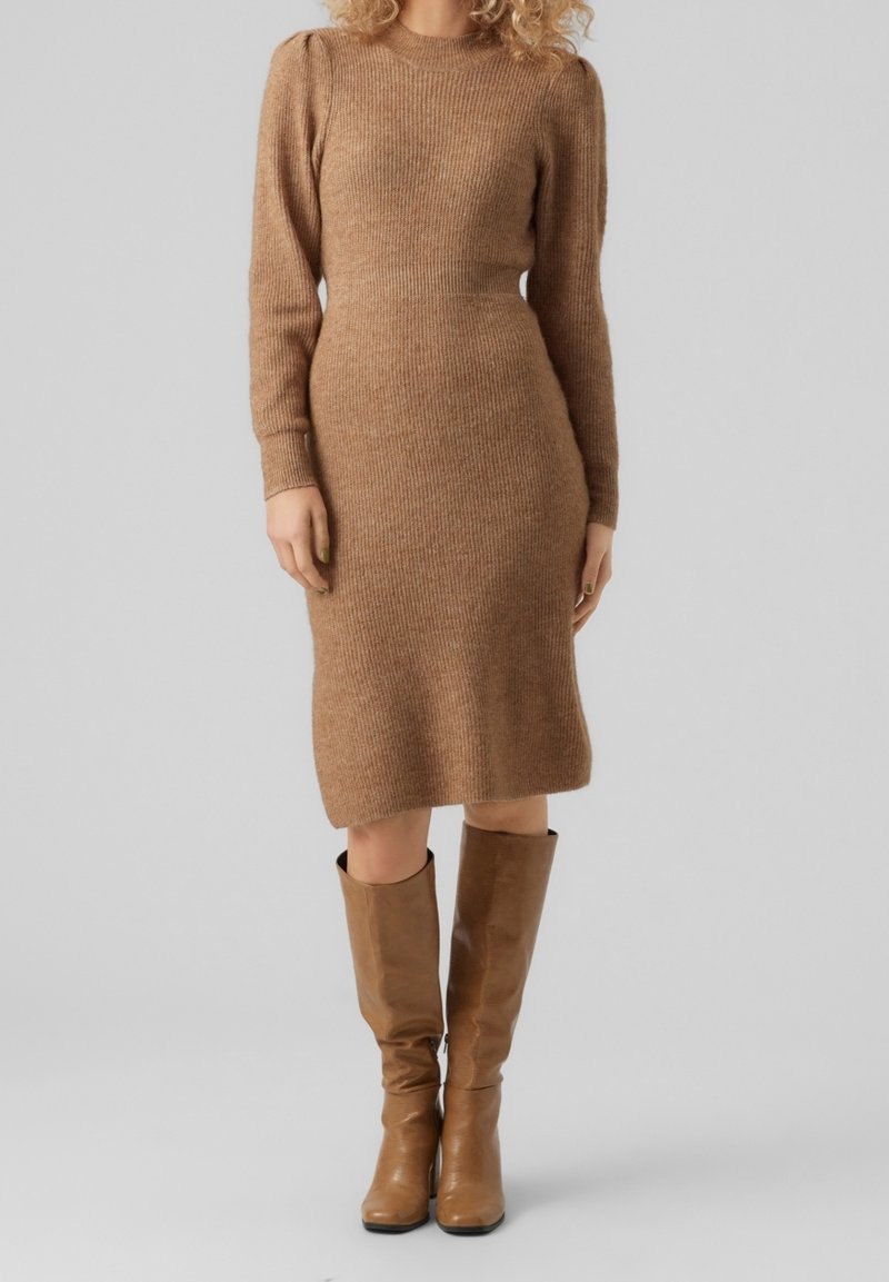 Femme portant une robe en maille marron à manches longues et longueur genou, associée à des bottes en cuir marron jusqu'aux genoux, debout devant un fond uni.