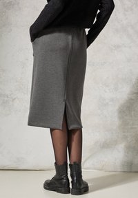 Grauer Jersey-Bleistiftrock mit einem seitlichen Schlitz, ausgestattet mit einem elastischen Bund und einer eleganten, glatten Textur. Getragen mit schwarzen Knöchelstiefeln.