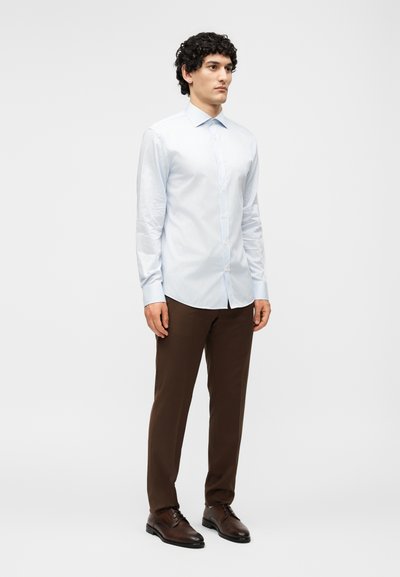 Άνδρας στέκεται φορώντας ένα ανοιχτό μπλε ριγέ πουκάμισο με μακριά μανίκια, σκουρόκαφέ παντελόνι και σκουρόκαφέ επίσημα παπούτσια ενάντια σε ένα απλό λευκό φόντο.