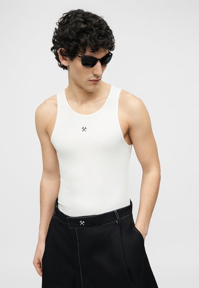 Άνδρας με σγουρά μαλλιά, φοράει μαύρα γυαλιά ηλίου, λευκό αμάνικο τοπ και μαύρο παντελόνι με ορατές ραφές, στέκεται με το ένα χέρι στην τσέπη.