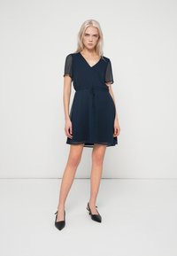 Ναυτικό μπλε φόρεμα με κοντά διαφανή μανίκια, V-λαιμόκοψη, μάζεμα στη μέση και φαρδύ φούστα. Συνδυασμένο με μαύρες γόβες με αιχμηρή μύτη.