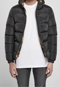 Czarna kurtka puchowa z kapturem wyłożonym w kamuflaż, zapinana z przodu na zamek, z elastycznymi mankietami i pikowanym wykończeniem. Nosi się ją na białej koszuli.