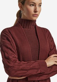 Mörkröd stickad tröja med hög ribbad krage och strukturerade kabelstickade ärmar. Mjuk tyg och avslappnad passform, med en enhetlig kropp.