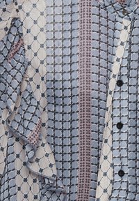Blusa multicolore con un motivo geometrico blu, rosa e crema, con leggere balze e bottoni neri lungo la parte anteriore.