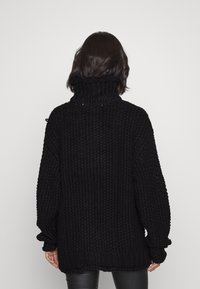 Schwarzer gestrickter Pullover mit hohem Stehkragen, lockerer Passform und gerippten Bündchen. Die Textur wirkt grob mit sichtbaren Nähdetails.